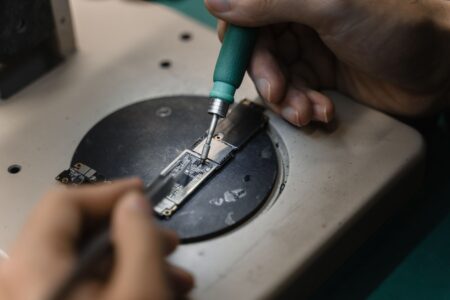 Hände, die ein Lötkolben auf einer Leiterplatte verwenden und präzise elektronische Reparatur in Innenräumen zeigen.