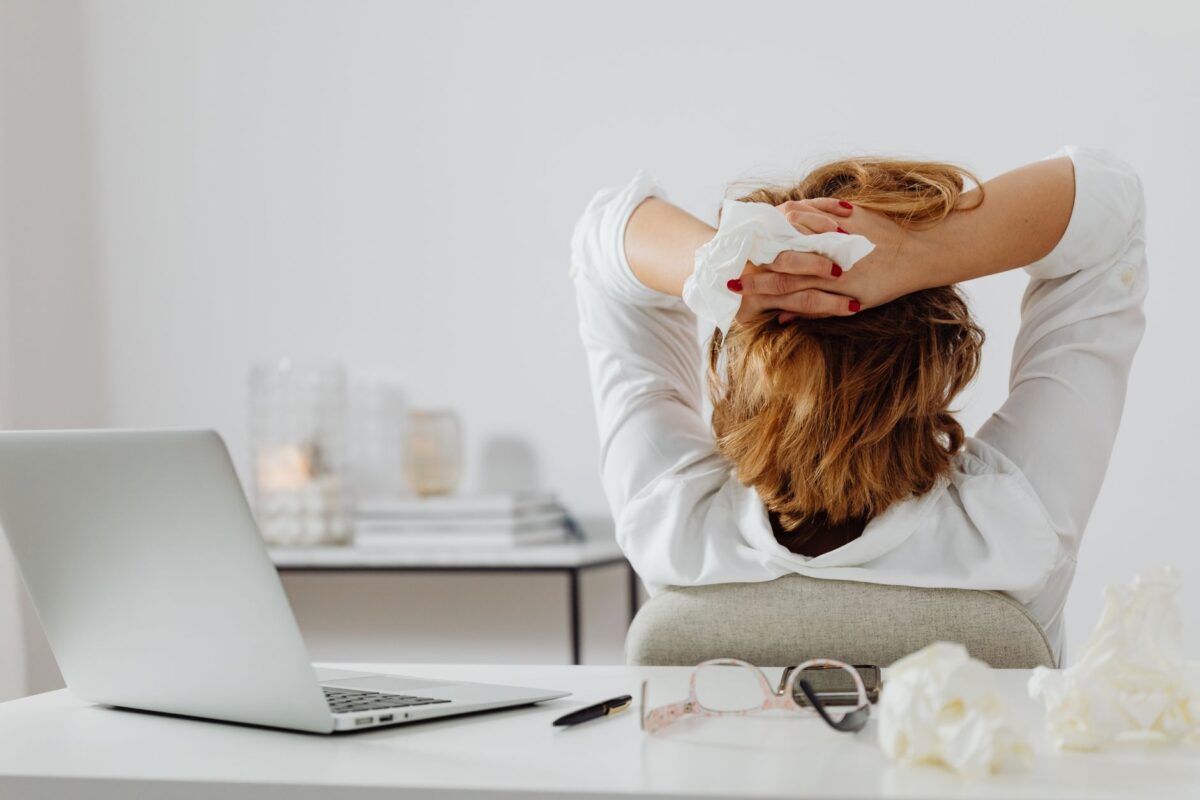 Ratgeber zum Thema Stressbewältigung: Strategien für den Alltag Eine erwachsene Frau in Weiß macht eine Pause an ihrem Schreibtisch, hält ein Taschentuch, während ein Laptop und eine