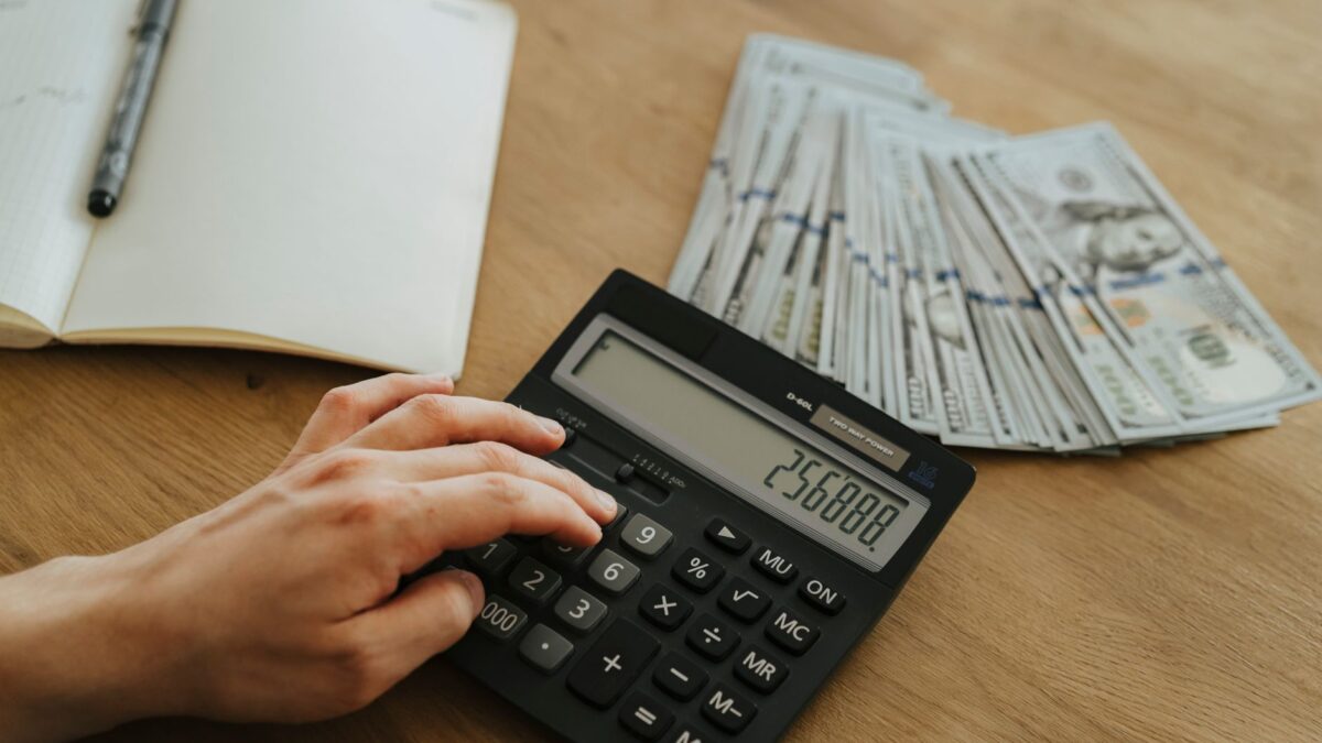 Eine Hand berechnet finanzielle Zahlen mit einem Taschenrechner, während nebenan auf einem Holztisch Geldscheine liegen.