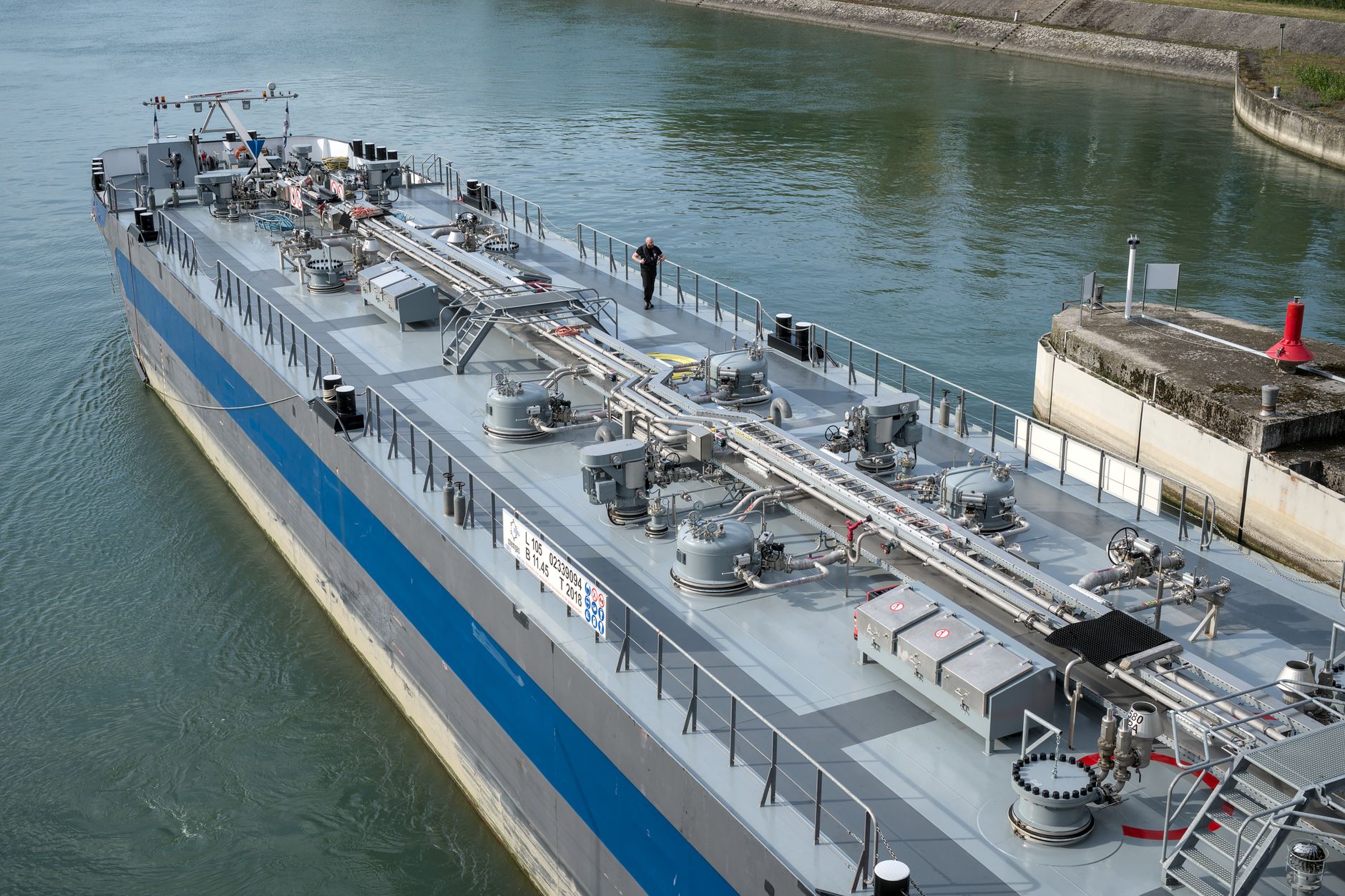Blick von oben auf ein graues Tankschiff mit Rohrleitungen und Tankdeckeln auf dem Deck, das auf ruhigem Wasser neben einer Kaimauer liegt.