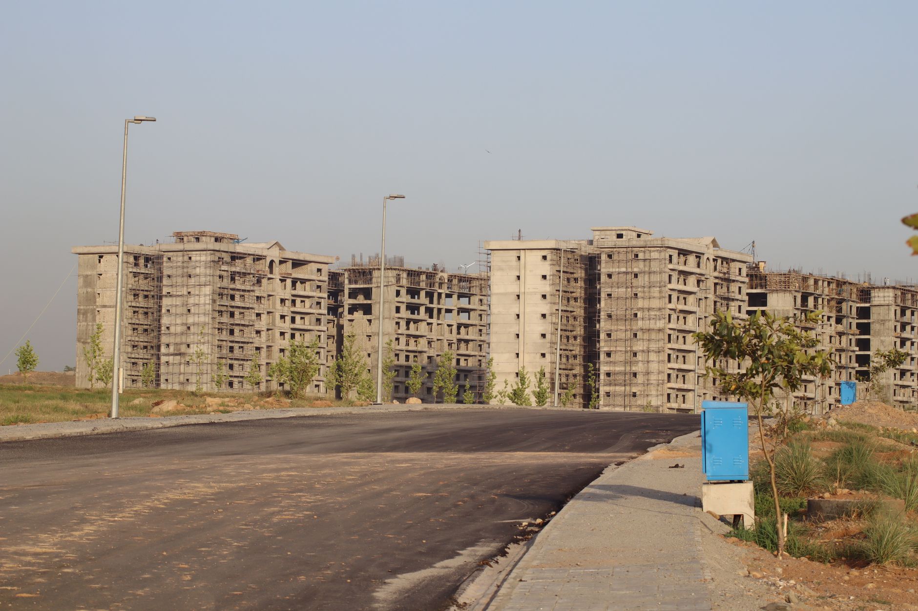 Unfertige Wohnblöcke im Betonrohbau mit Gerüsten hinter einer leeren, frisch asphaltierten Straße, davor Gehweg, Laternen und junge Bäume